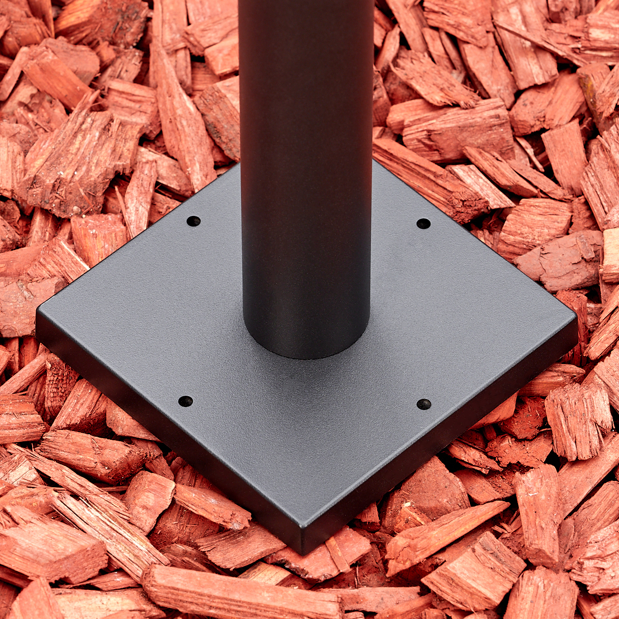 Ein schwarzer Metallpfosten auf einer quadratischen Basis mit vier Löchern, auf roten Holzspänen. Der Pfosten ist zentriert.