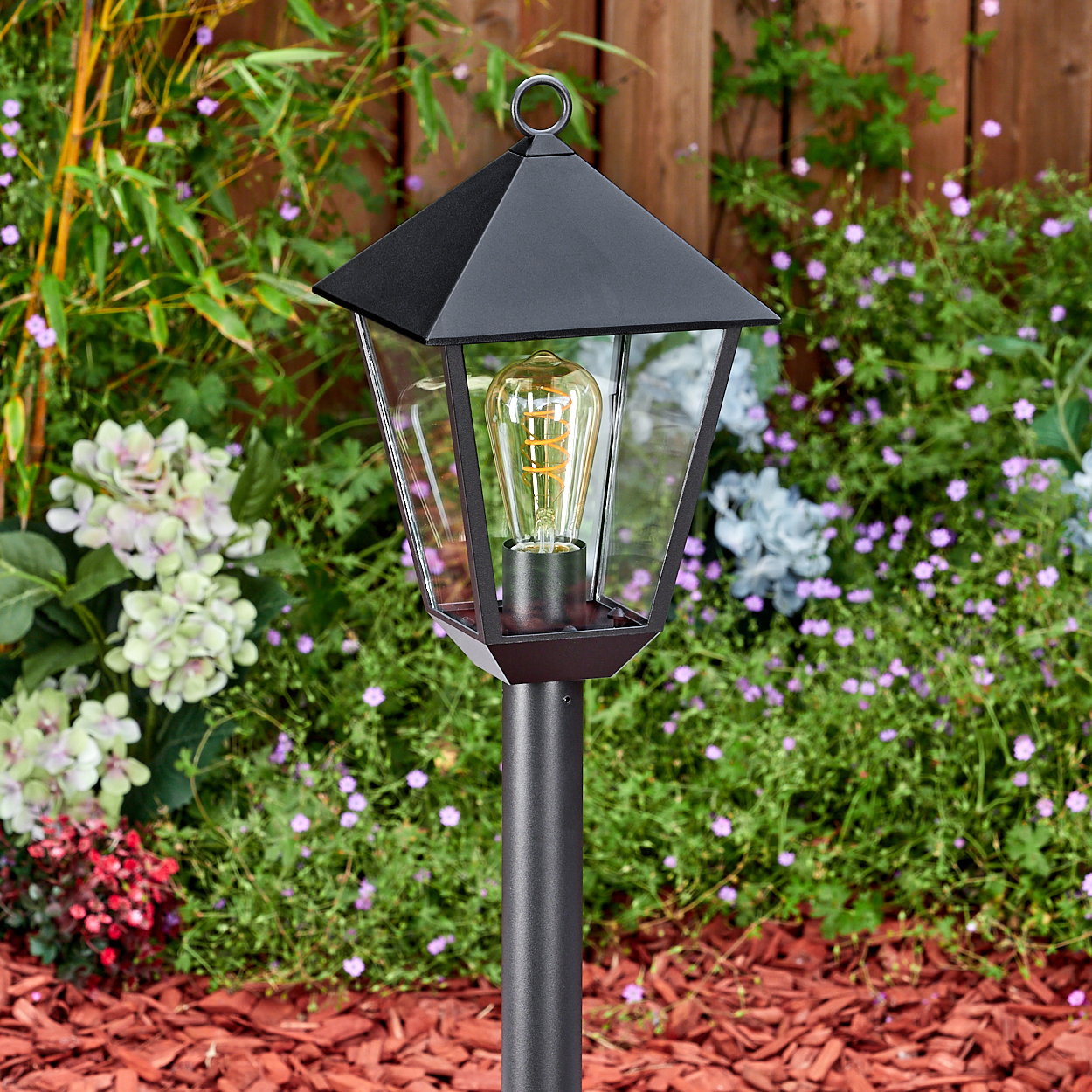Eine schwarze Außenlampe mit sichtbarer Glühbirne steht in einem Garten mit Blumen und einem Holzzaun im Hintergrund.