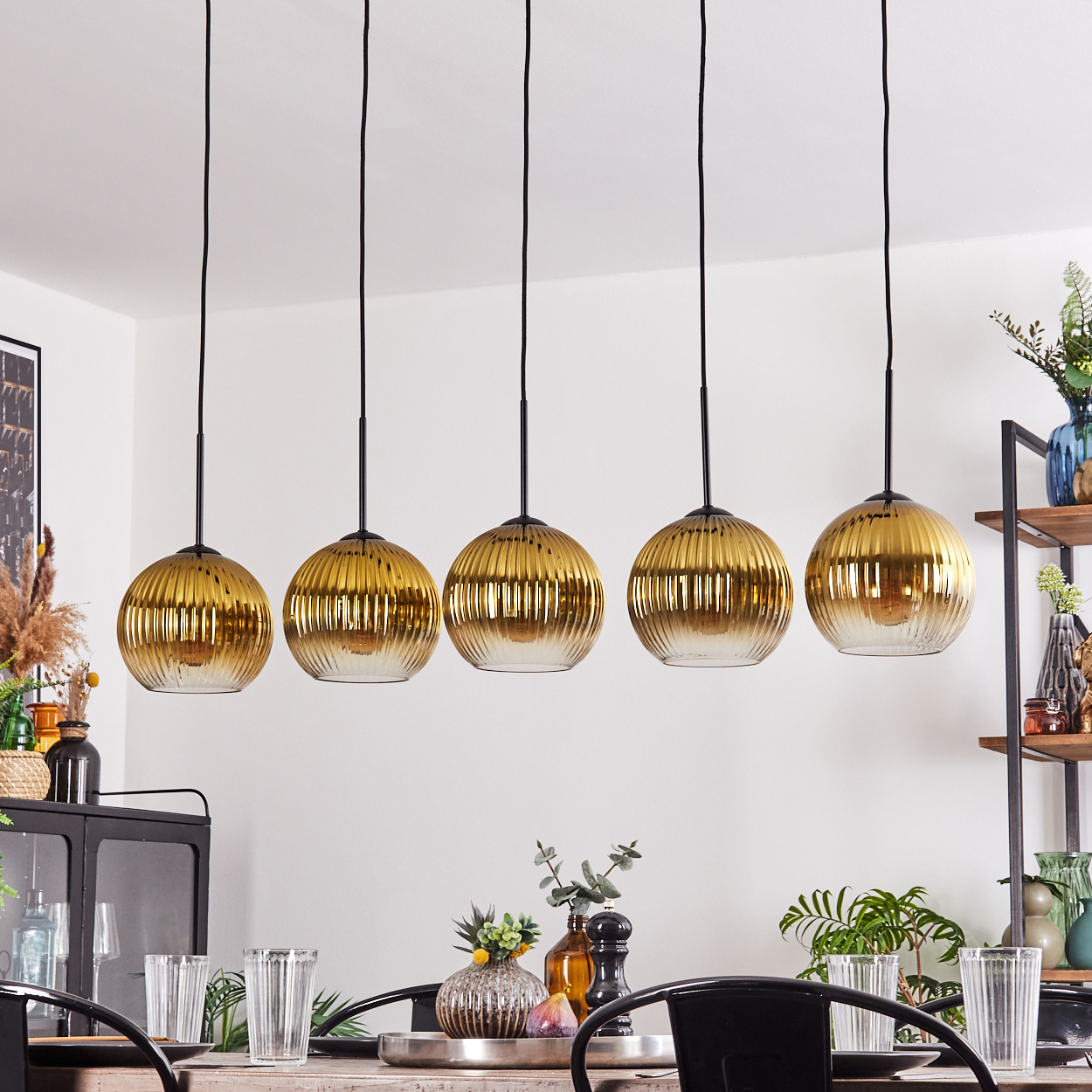 Fünf goldene Hängelampen hängen über einem Tisch mit Gläsern und Dekorationen. Im Hintergrund ist ein Regal mit Vasen zu sehen.