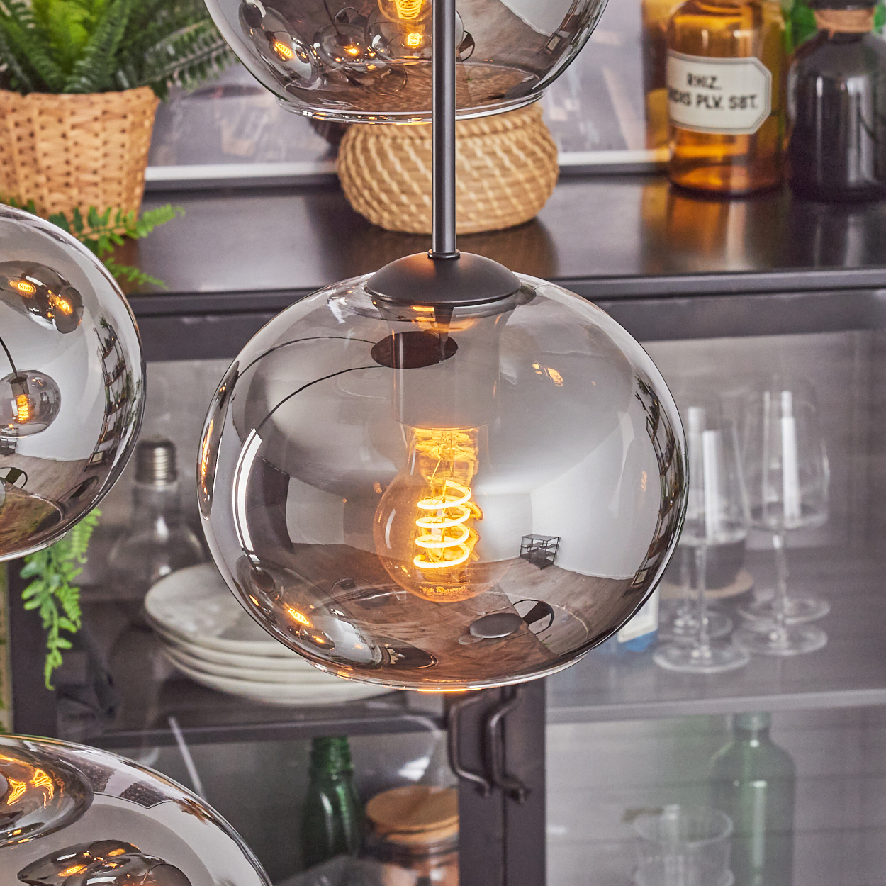 Eine moderne Hängelampe mit drei Glaskugeln, jede mit einer Wendelfadenlampe. Im Hintergrund Pflanzen, Geschirr und Flaschen.