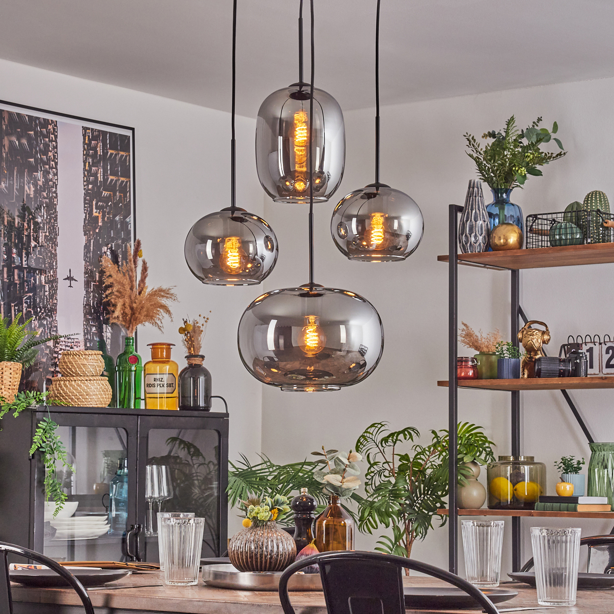 Esszimmer mit Hängelampen, Pflanzen und einem gedeckten Tisch. Dekorative Gegenstände auf Regalen und ein schwarzer Schrank sind sichtbar.