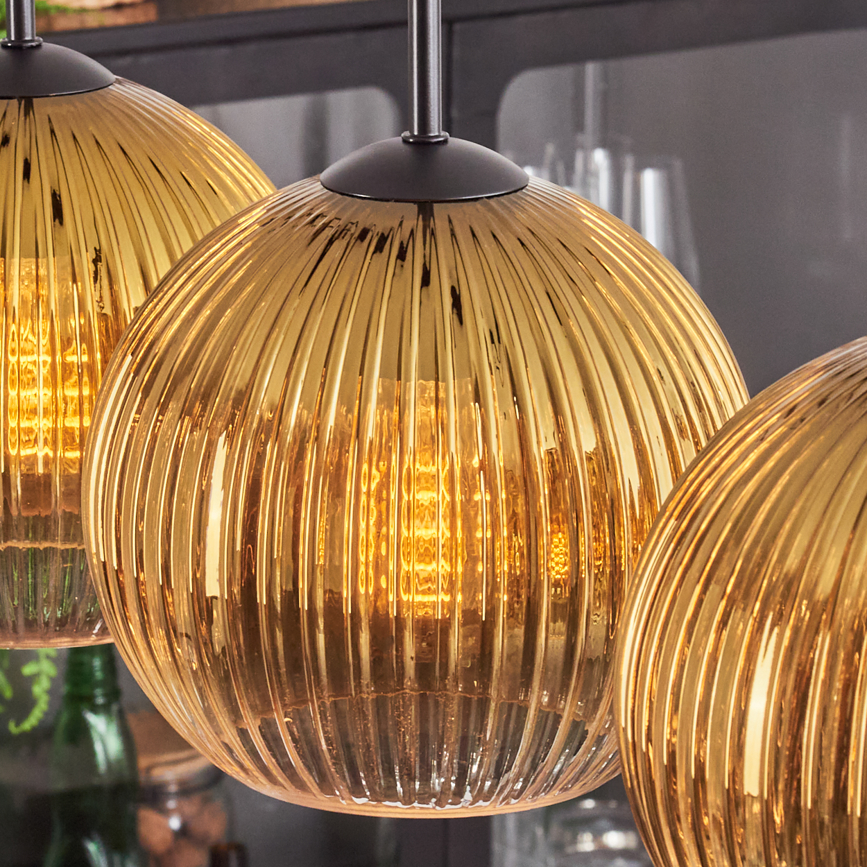 Drei goldene, gerippte Glas-Pendelleuchten mit sichtbaren Glühbirnen. Hintergrund zeigt verschwommene dunkle Oberflächen und eine grüne Flasche.