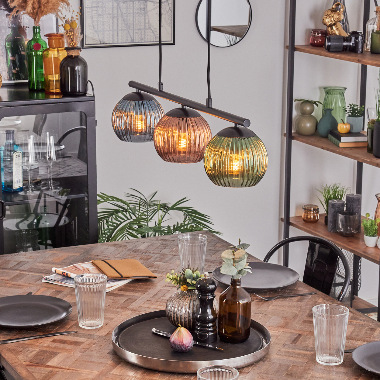 Esszimmer-Szene mit einem Holztisch, der für ein Essen gedeckt ist. Drei farbige Pendelleuchten darüber, Regal im Hintergrund mit Dekoration.