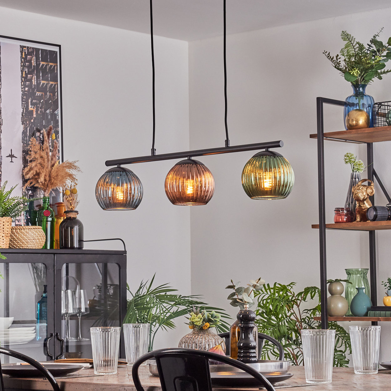 Esszimmer mit dreifach-Pendelleuchte. Farbig getönte Glasschirme hängen über einem gedeckten Tisch. Regale mit Vasen rechts.