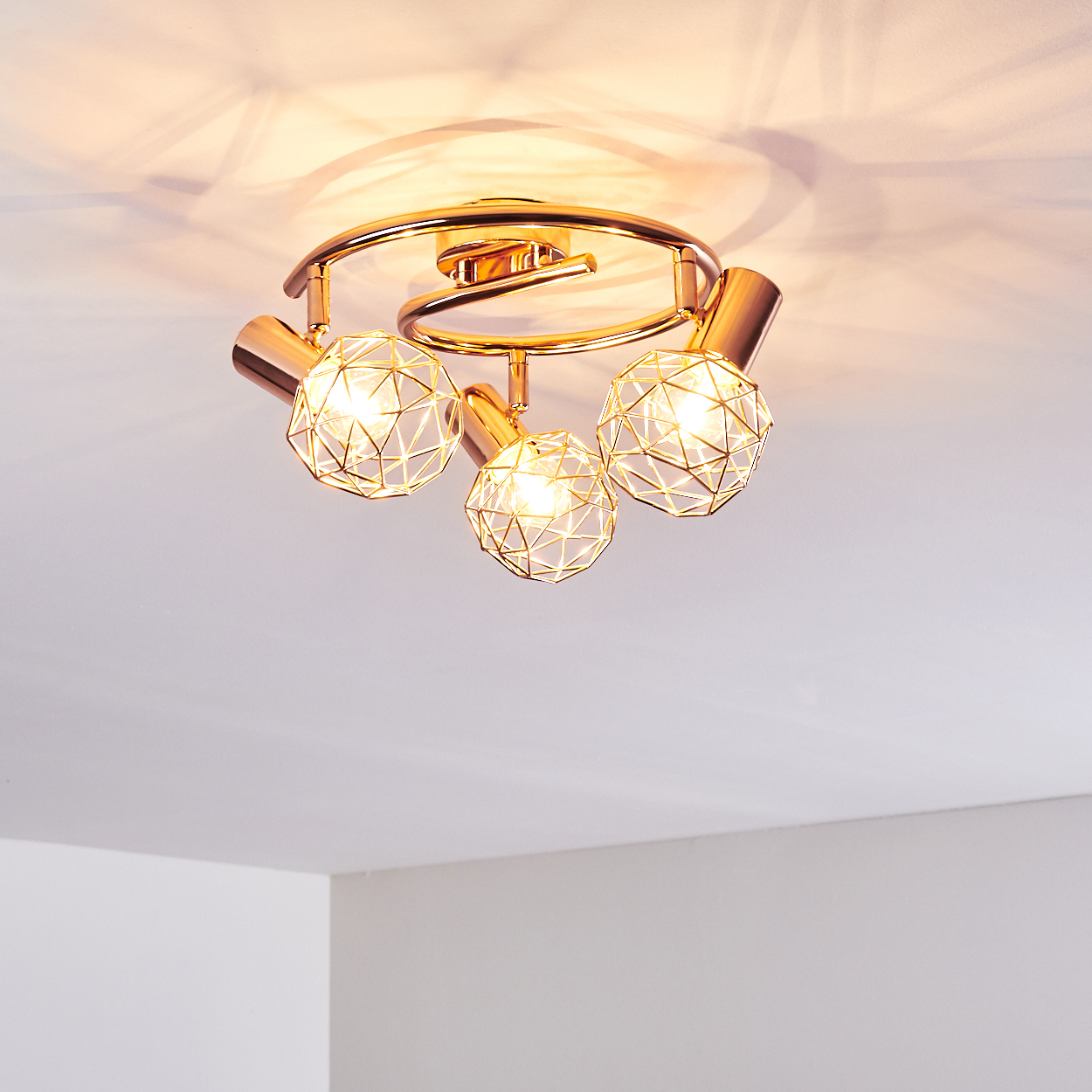 Eine Deckenleuchte mit drei kugelförmigen Käfigleuchten, goldene Farbe. Die Decke ist weiß.
