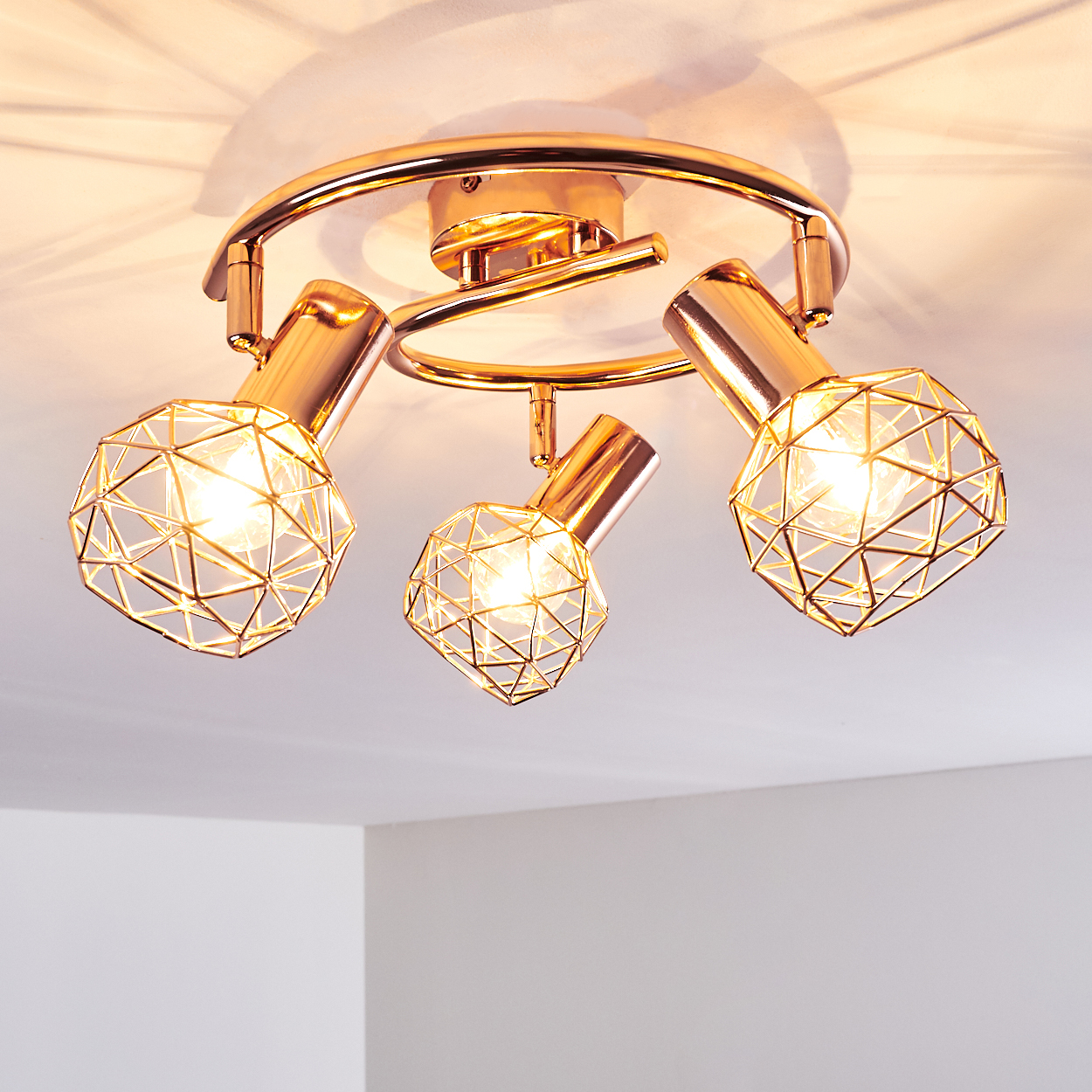 Eine goldene Deckenleuchte mit drei verstellbaren Strahlern, die jeweils von einem geometrischen Drahtrahmen umgeben sind, vor einer weißen Decke.