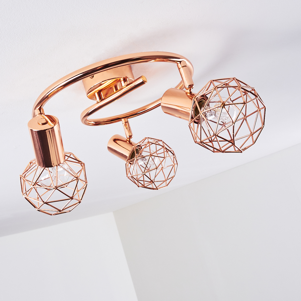 Eine roségoldene Deckenleuchte mit drei geometrischen Käfigbirnen. Die Leuchte ist an einer weißen Decke montiert.
