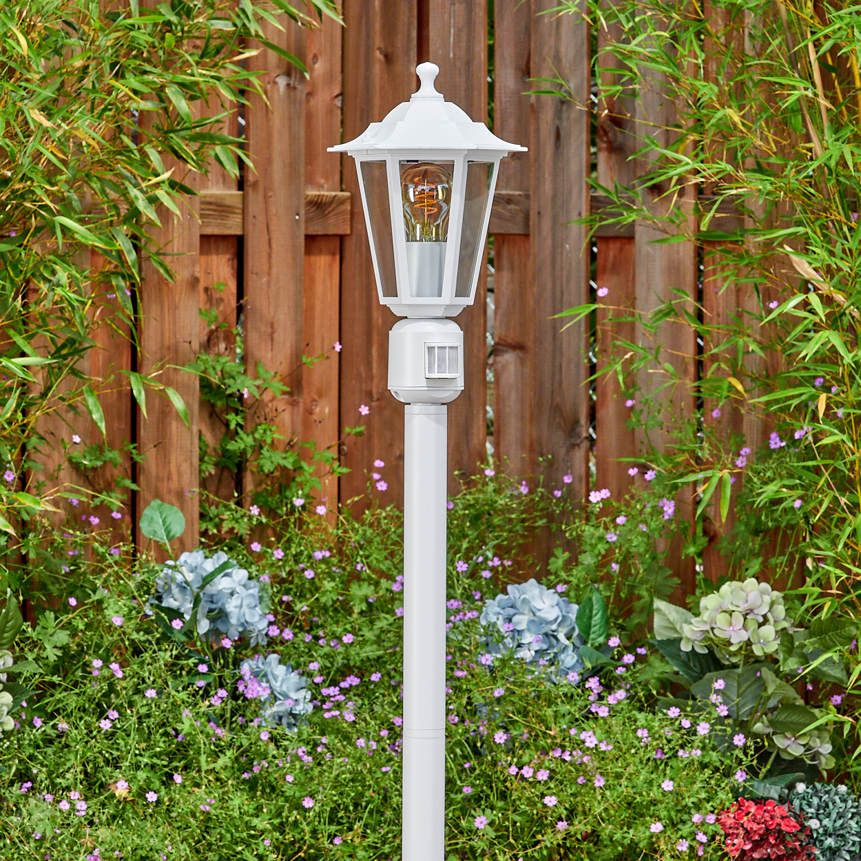 Eine weiße Außenlampe steht in einem Garten. Bambus und Blumen umgeben sie. Im Hintergrund ist ein Holzzaun.