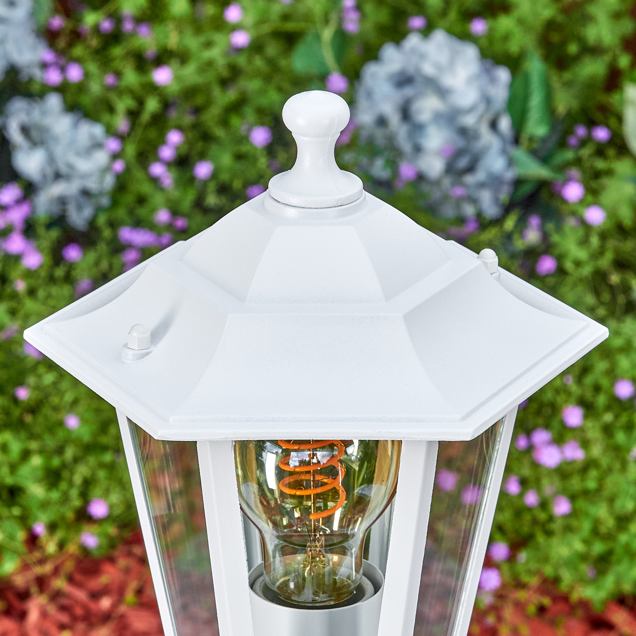 Weiße Außenlampe mit sichtbarer Glühbirne, umgeben von grünem Laub und lila Blüten. Rindenmulch im Hintergrund.