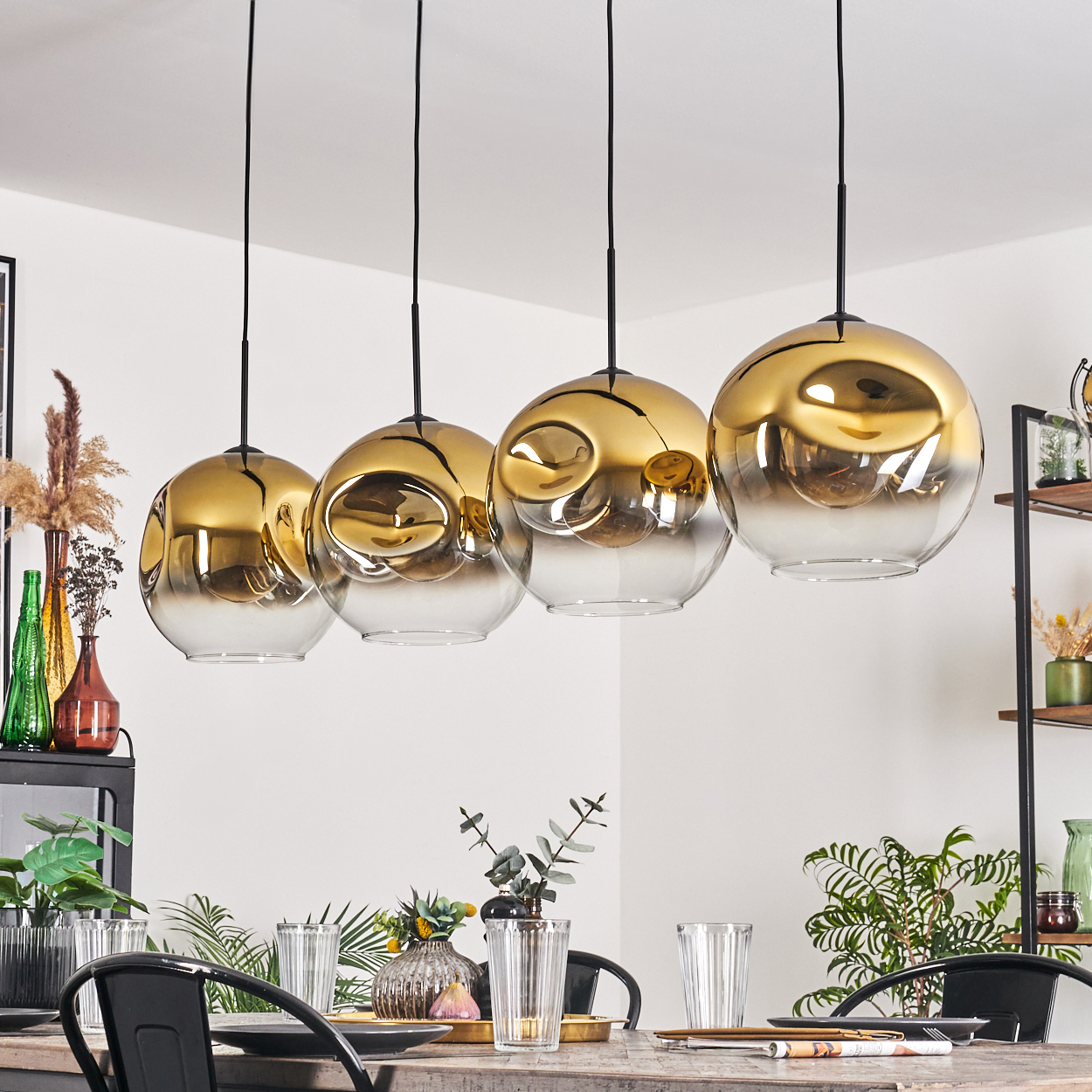 Vier kugelförmige gold- und gläserne Pendelleuchten hängen über einem Esstisch, der für eine Mahlzeit gedeckt ist. Pflanzen und Dekorationen sind sichtbar.
