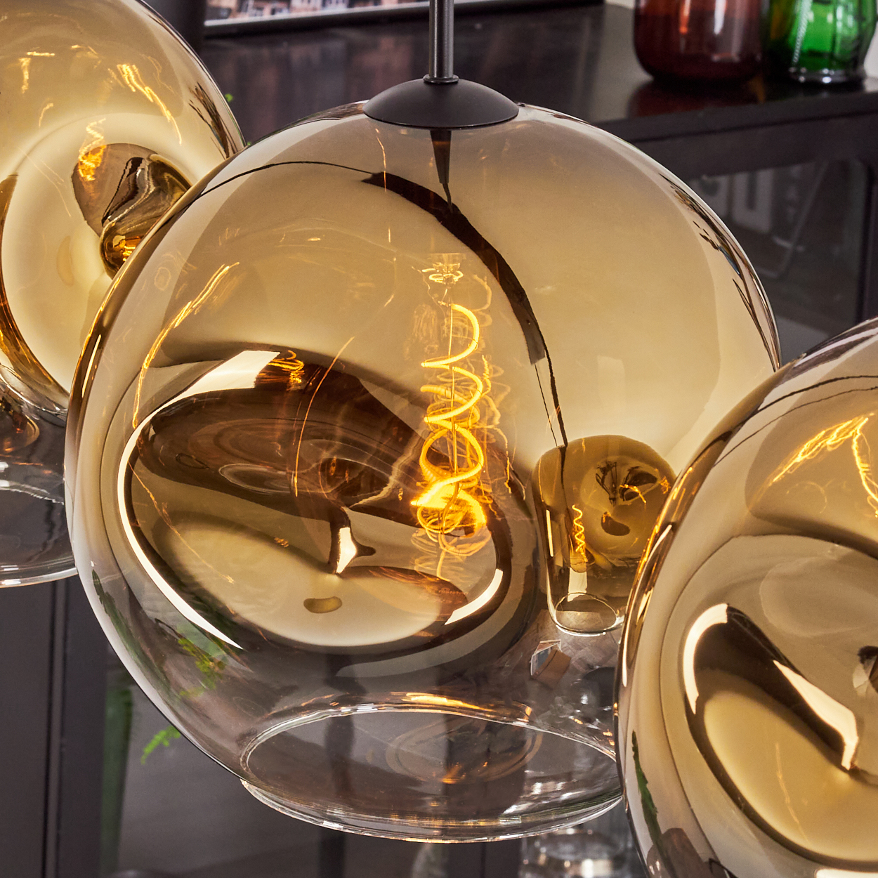 Drei goldfarbene, kugelförmige Pendelleuchten mit sichtbaren Glühfäden. Licht reflektiert im Glas. Dunkler Hintergrund.