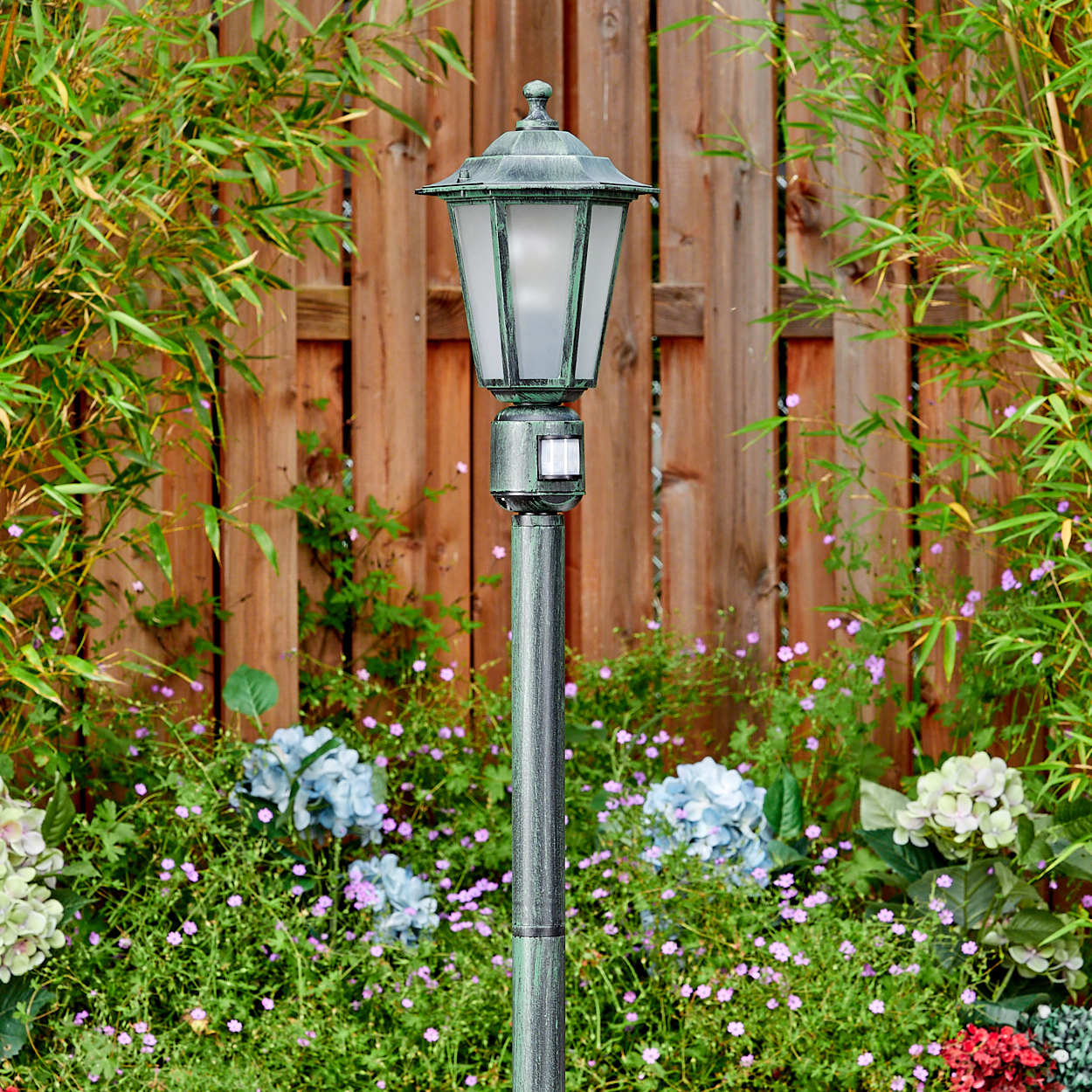 Eine grüne Laterne in einem Garten, mit Holzzaun und Bambus im Hintergrund und Blumen im Vordergrund.