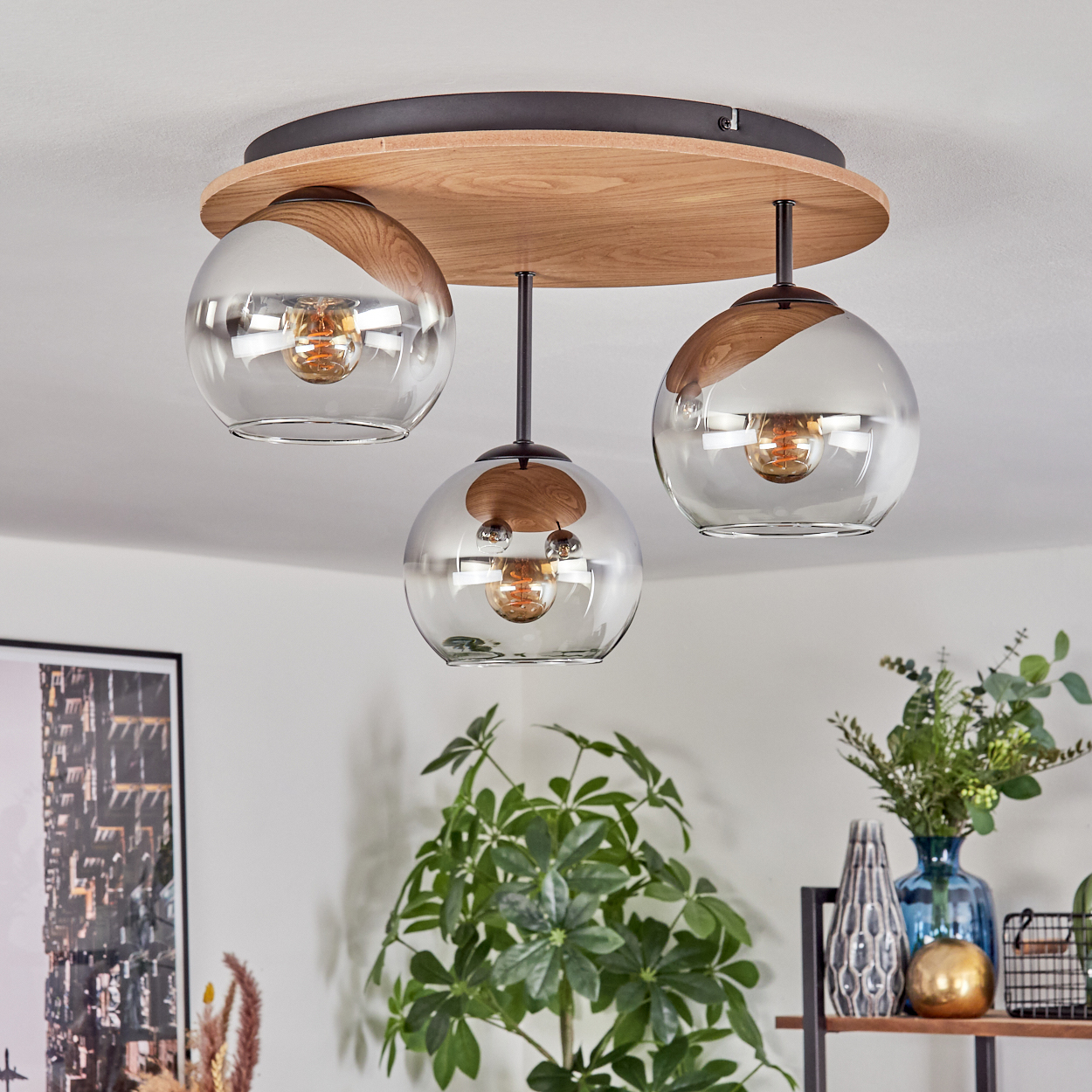 Deckenlampe mit drei Glaskugeln. Akzente aus Holz und schwarzem Metall. Pflanzen und Dekorationsartikel sind im Hintergrund sichtbar.