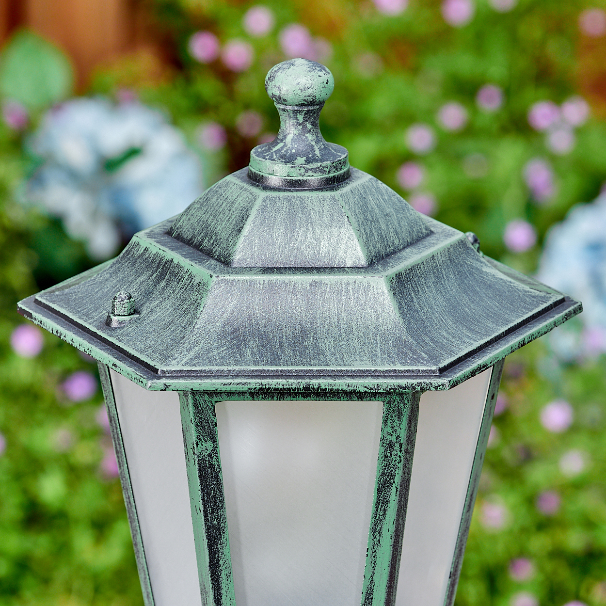 Eine grün-schwarze Außenlampe. Die Lampe hat eine sechseckige Oberseite und Milchglas. Verschwommene Blumen sind im Hintergrund.