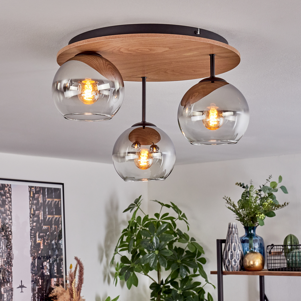 Moderne Deckenlampe mit drei Glaskugeln. Die Lampe hängt an einem Holzsockel. Pflanzen und Dekoration im Hintergrund.