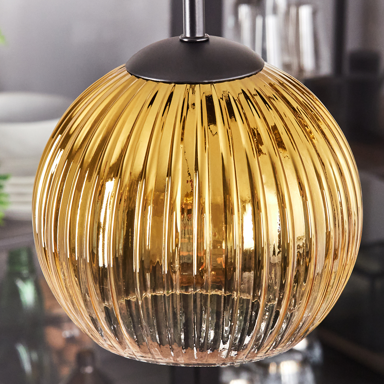 Nahaufnahme einer goldenen, geriffelten Glas-Hängeleuchte. Sie hängt an einer schwarzen Halterung, vor verschwommenem Hintergrund.