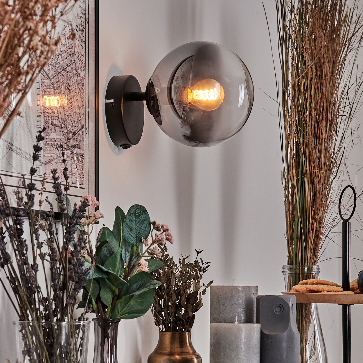 Eine moderne Wandleuchte mit Kugelschirm. Dekorative Elemente sind Trockenblumen, Kerzen und ein Kartendruck.