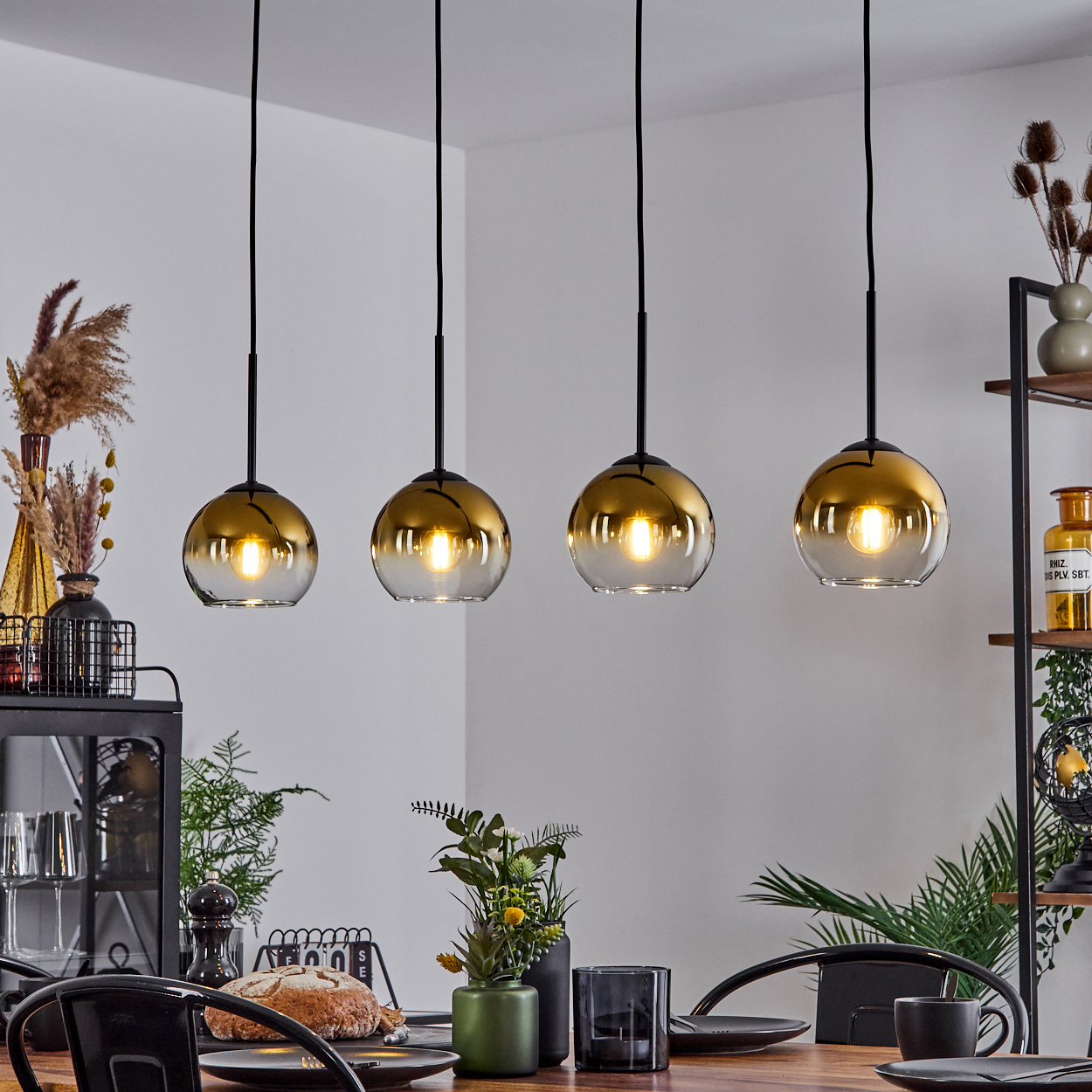 Vier goldfarbene Glas-Hängelampen hängen über einem Holztisch, der für eine Mahlzeit gedeckt ist. Im Hintergrund Regale und Pflanzen.