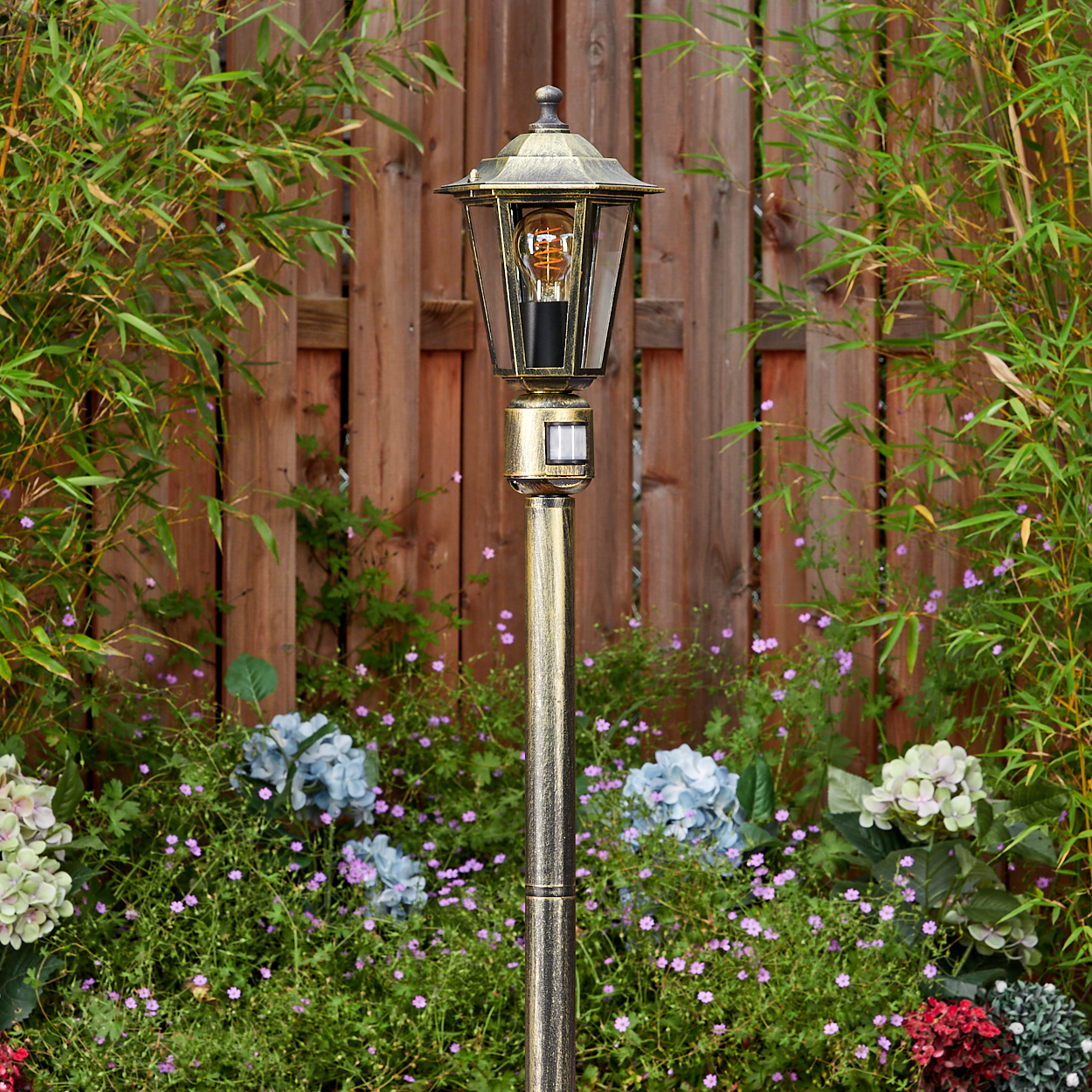 Eine goldene Außenlampe steht in einem Garten. Der Hintergrund zeigt einen Holzzaun und verschiedene grüne Pflanzen und Blumen.