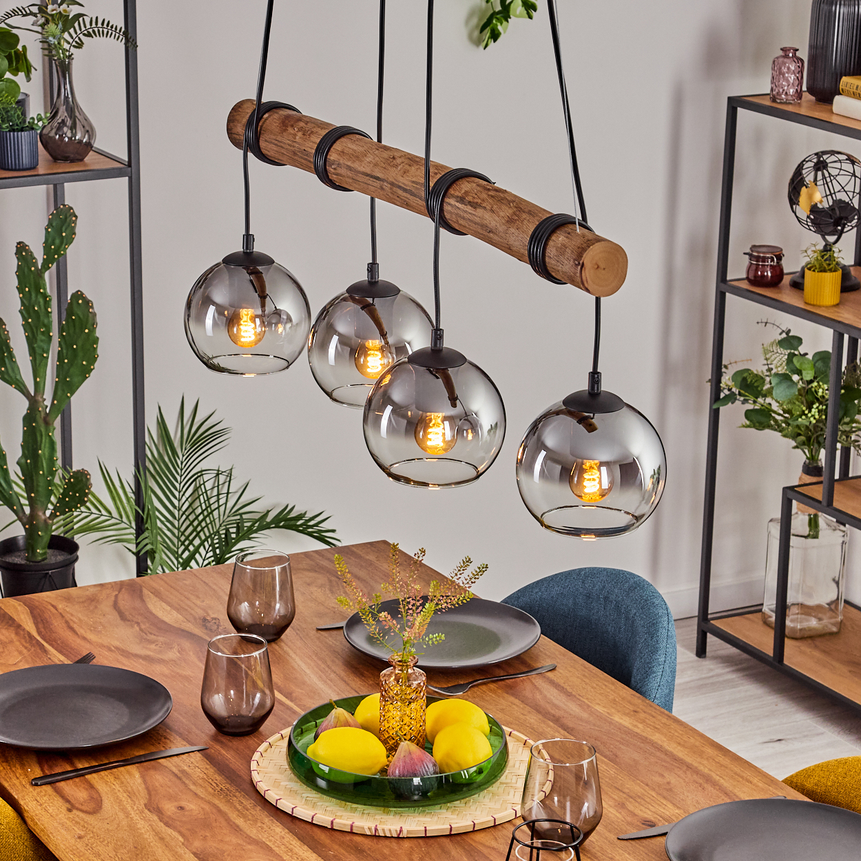 Esstisch mit Hängelampen. Vier Glaskugelleuchten hängen über einem Holztisch mit Früchten und Tellern. Regale im Hintergrund.