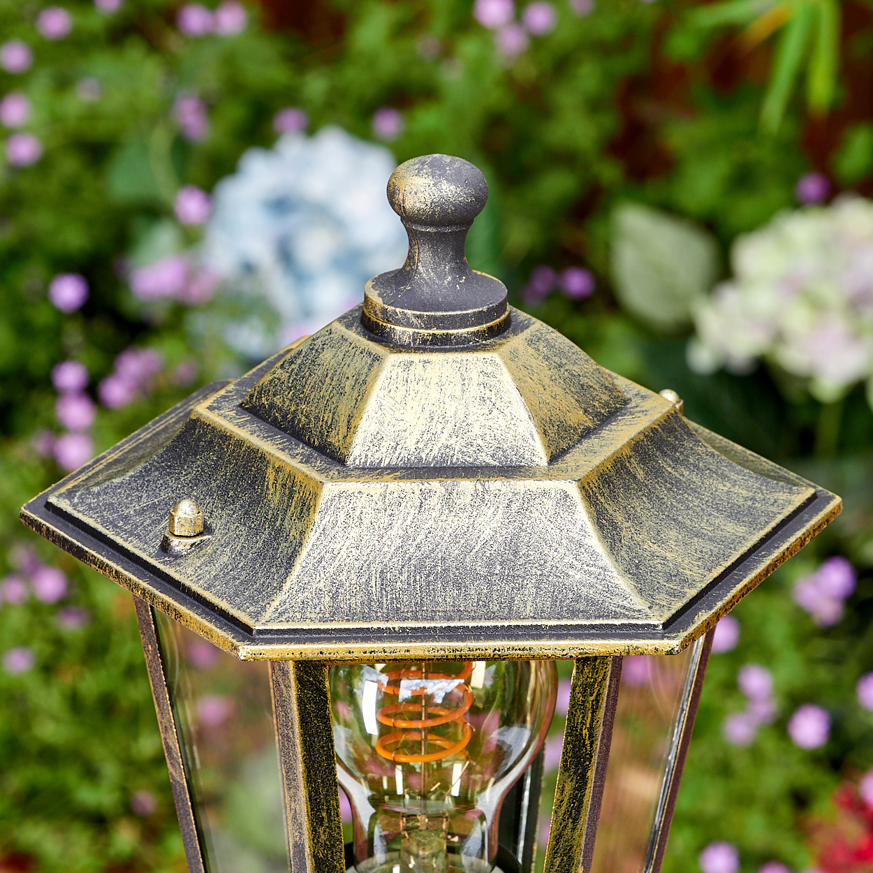 Nahaufnahme einer dekorativen Außenlampe mit goldenem Dach und freiliegender Glühbirne. Der Hintergrund ist verschwommenes grünes Laub und Blumen.