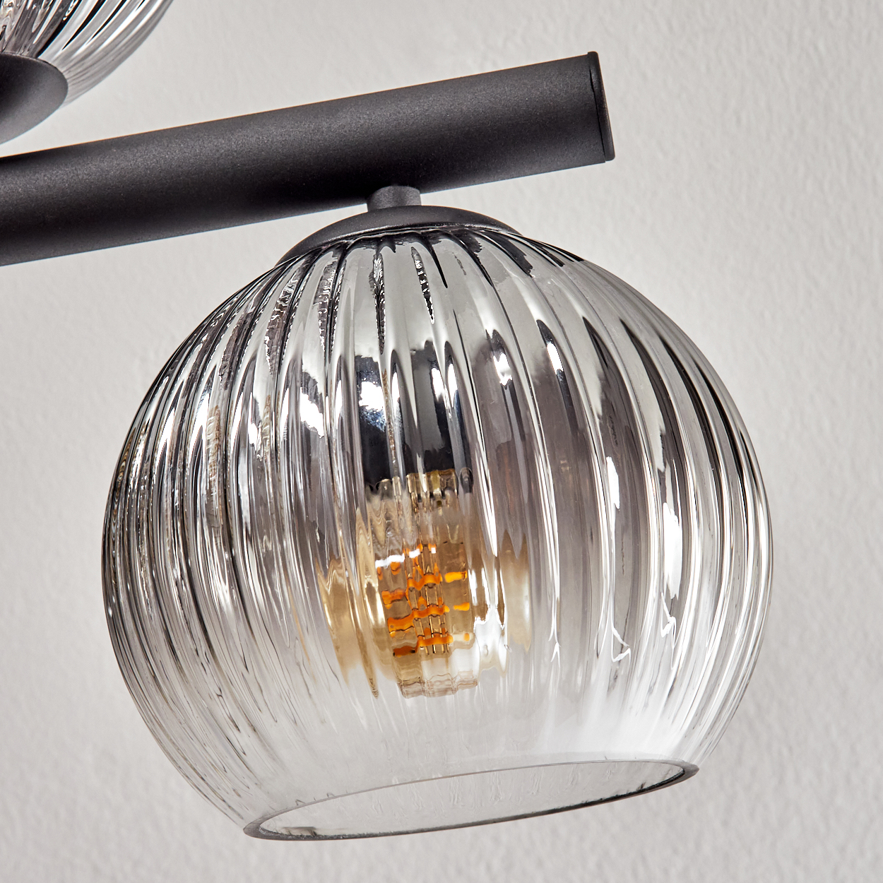 Nahaufnahme einer Leuchte. Sie hat einen gerippten Glasschirm und eine sichtbare Glühbirne. Die Leuchte befindet sich vor einer weißen Wand.