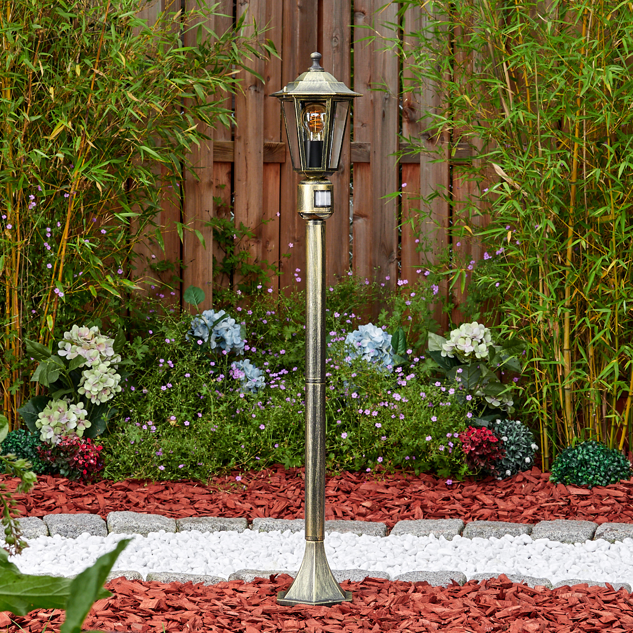 Eine goldene Laterne steht in einem Garten mit Bambus, Blumen und einem hölzernen Zaun im Hintergrund.