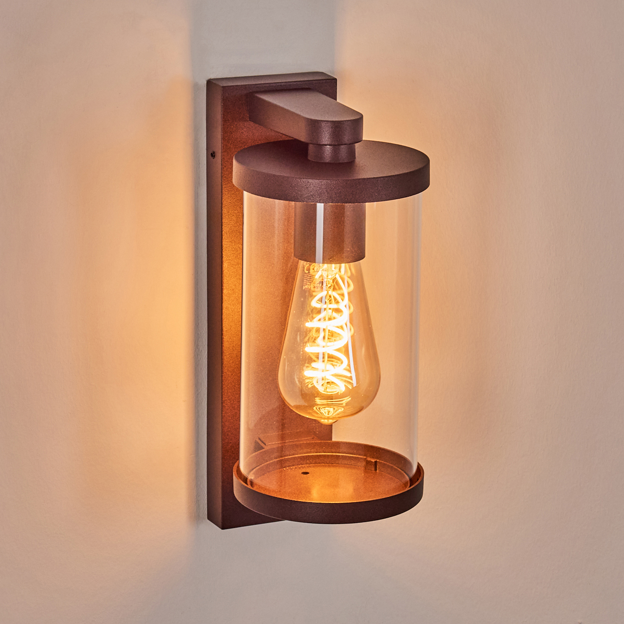 Eine bronzefarbene Wandlampe mit Glaszylinder. Im Inneren befindet sich eine Wendelglühbirne, die ein warmes Licht auf eine beige Wand wirft.