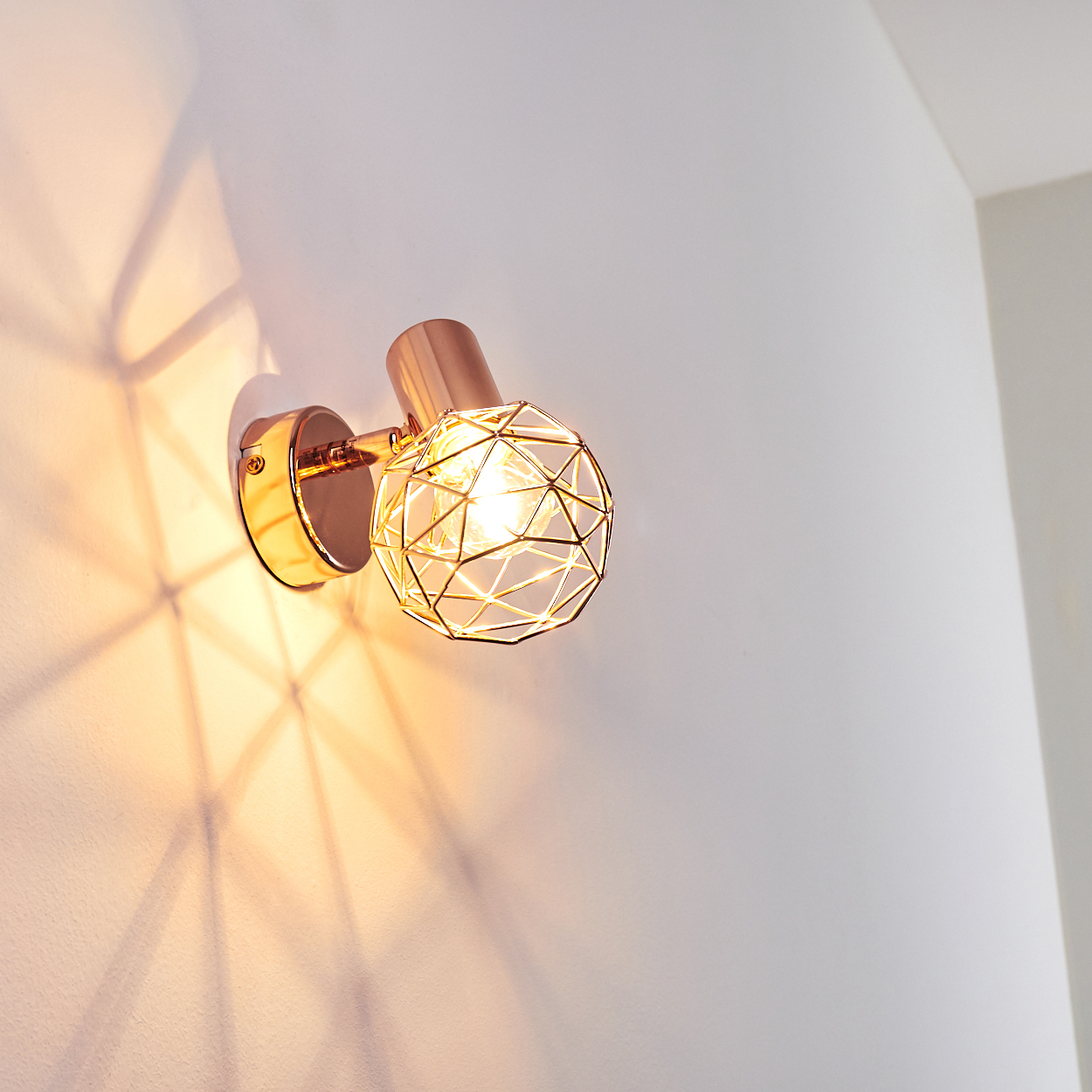 Eine goldene Wandlampe mit geometrischem Käfigdesign. Sie wirft einen Schatten auf die weiße Wand. Die Lampe ist an.