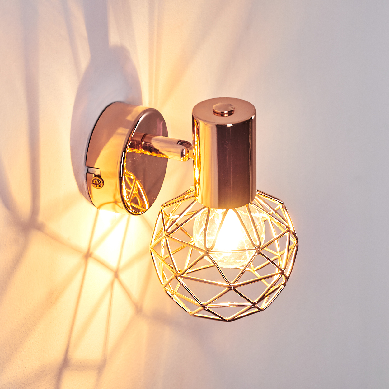 Eine roségoldene Wandleuchte mit geometrischem Käfigschirm. Das Licht wirft Schatten auf die weiße Wand.
