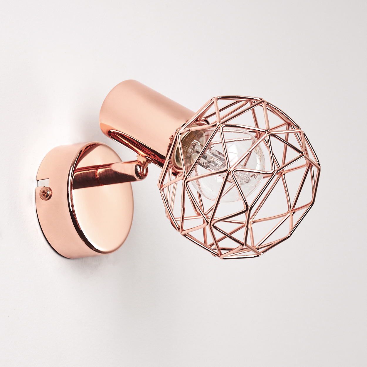 Eine roségoldene Wandleuchte mit geometrischem Schirm ist an einer weißen Wand befestigt. Die Lampe hat eine runde Basis und einen zylindrischen Arm.