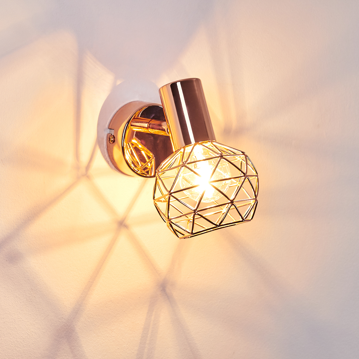 Eine goldene Wandleuchte mit geometrischem Käfigdesign, an einer weißen Wand. Die Lampe wirft ein gemustertes Licht.