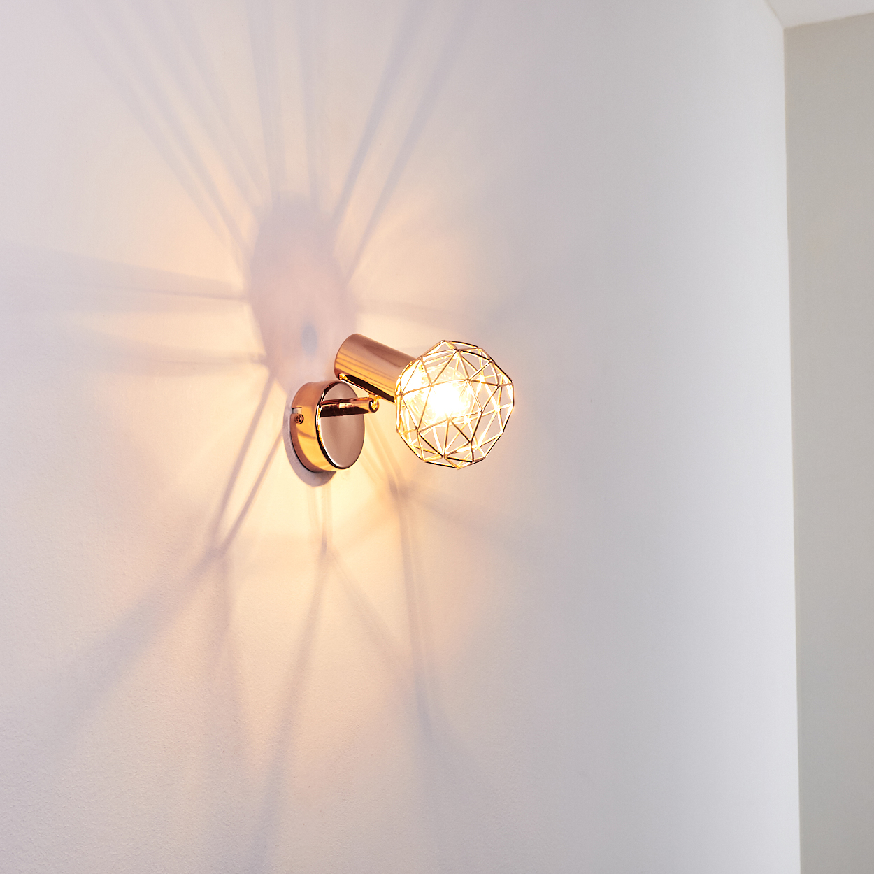 Eine goldene Wandlampe mit geometrischem Schirm ist an einer weißen Wand befestigt und wirft ein Lichtmuster.