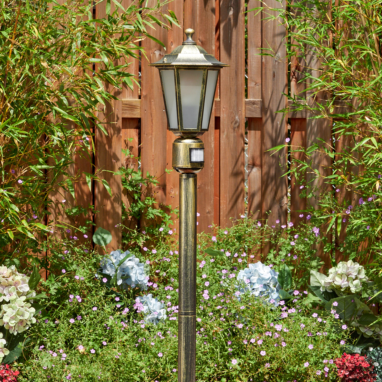Eine dekorative Außenlampe steht in einem Garten mit einem hölzernen Zaun im Hintergrund und verschiedenen Pflanzen und Blumen.