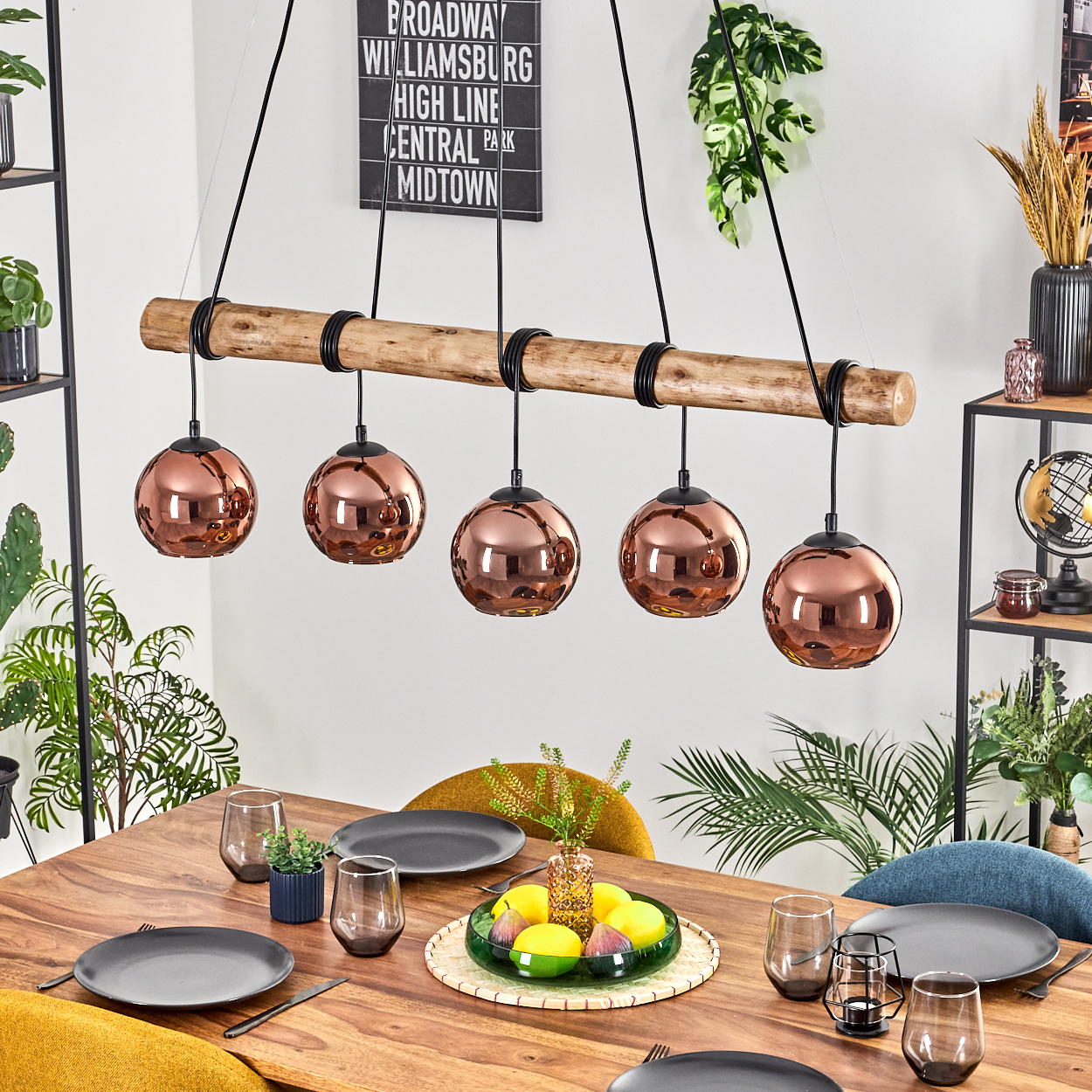 Esszimmer mit Holztisch, Kupfer-Hängelampen und einer Obstschale. Im Hintergrund Pflanzen, ein Regal und ein Wandzeichen.