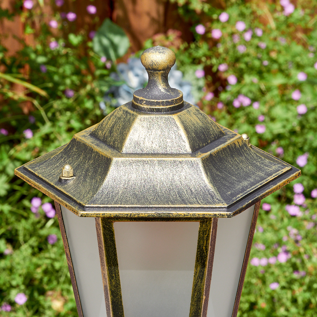 Nahaufnahme einer dekorativen Außenlampe mit Gold- und Schwarzfinish vor dem Hintergrund grüner Pflanzen und lila Blüten.