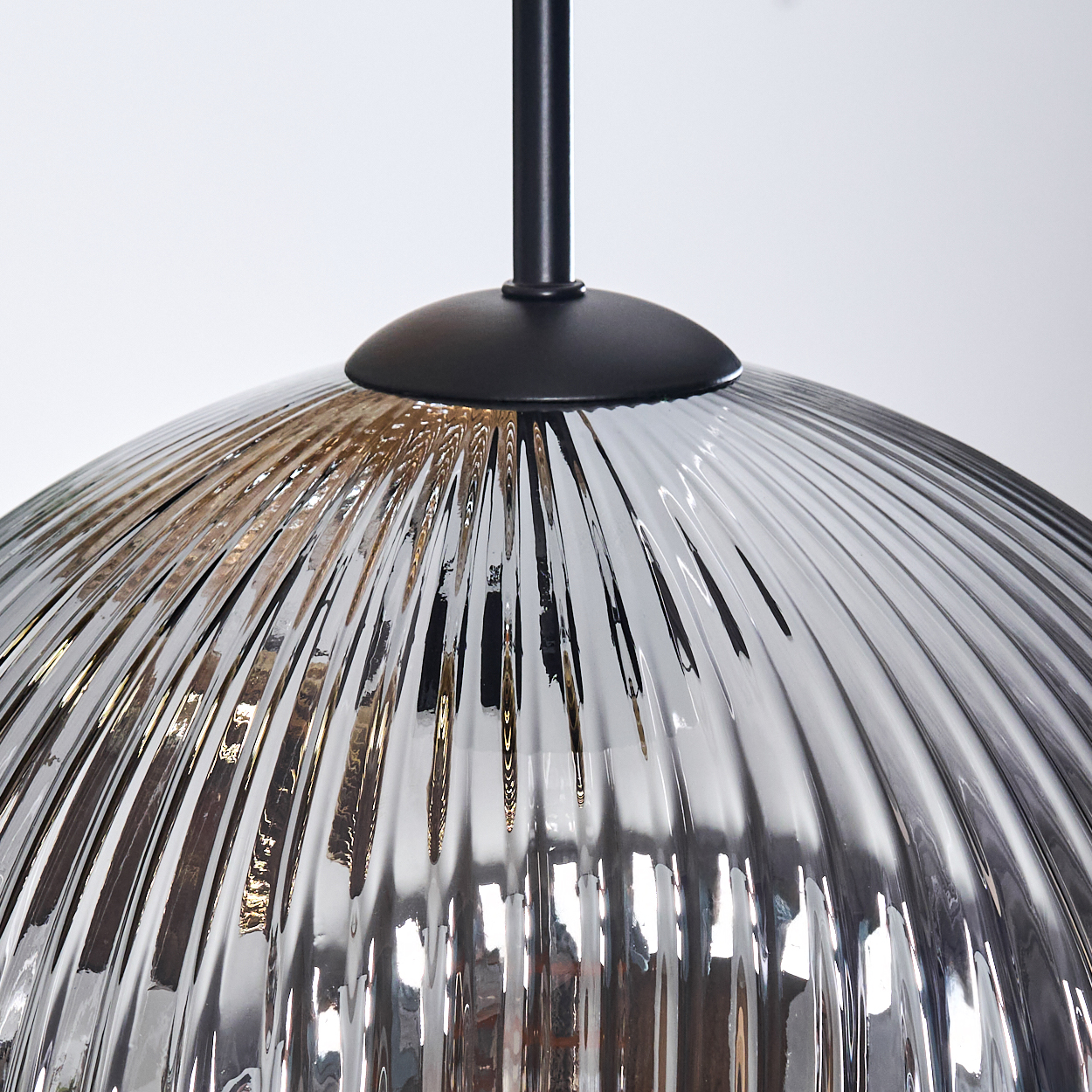 Nahaufnahme einer Glas-Hängelampe. Die Lampe hat einen gerippten, transparenten Glasschirm und eine schwarze Metallbefestigung vor weißem Hintergrund.