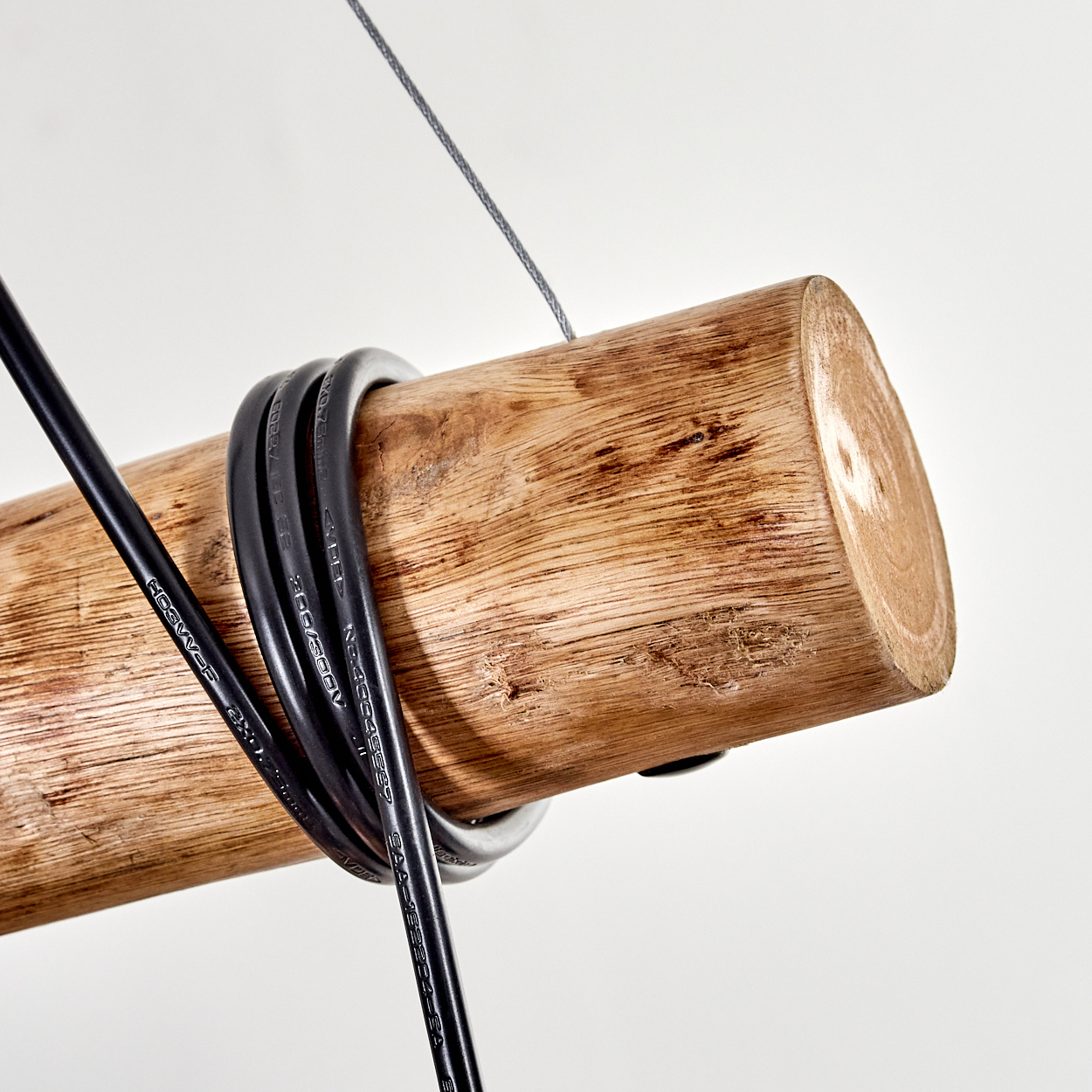 Ein Holzstamm mit schwarzen Kabeln. Ein grauer Draht hängt ihn vor weißem Hintergrund auf.