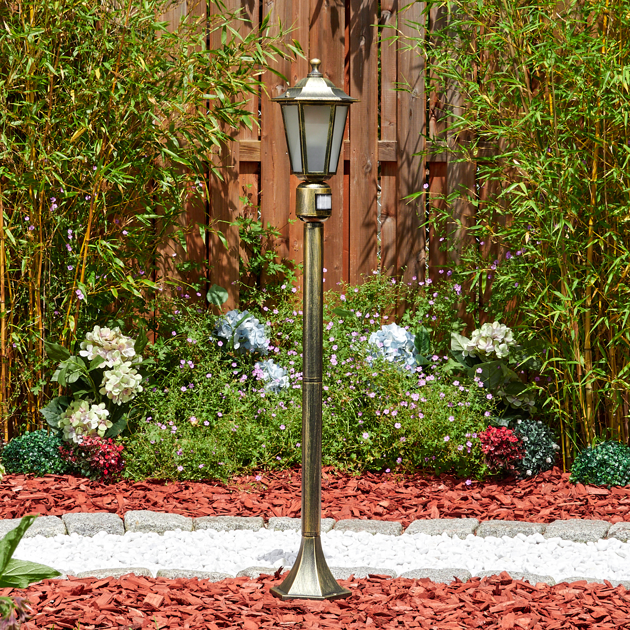 Eine bronzefarbene Straßenlaterne steht in einem Garten. Bambus, Blumen und ein Holzzaun befinden sich im Hintergrund.