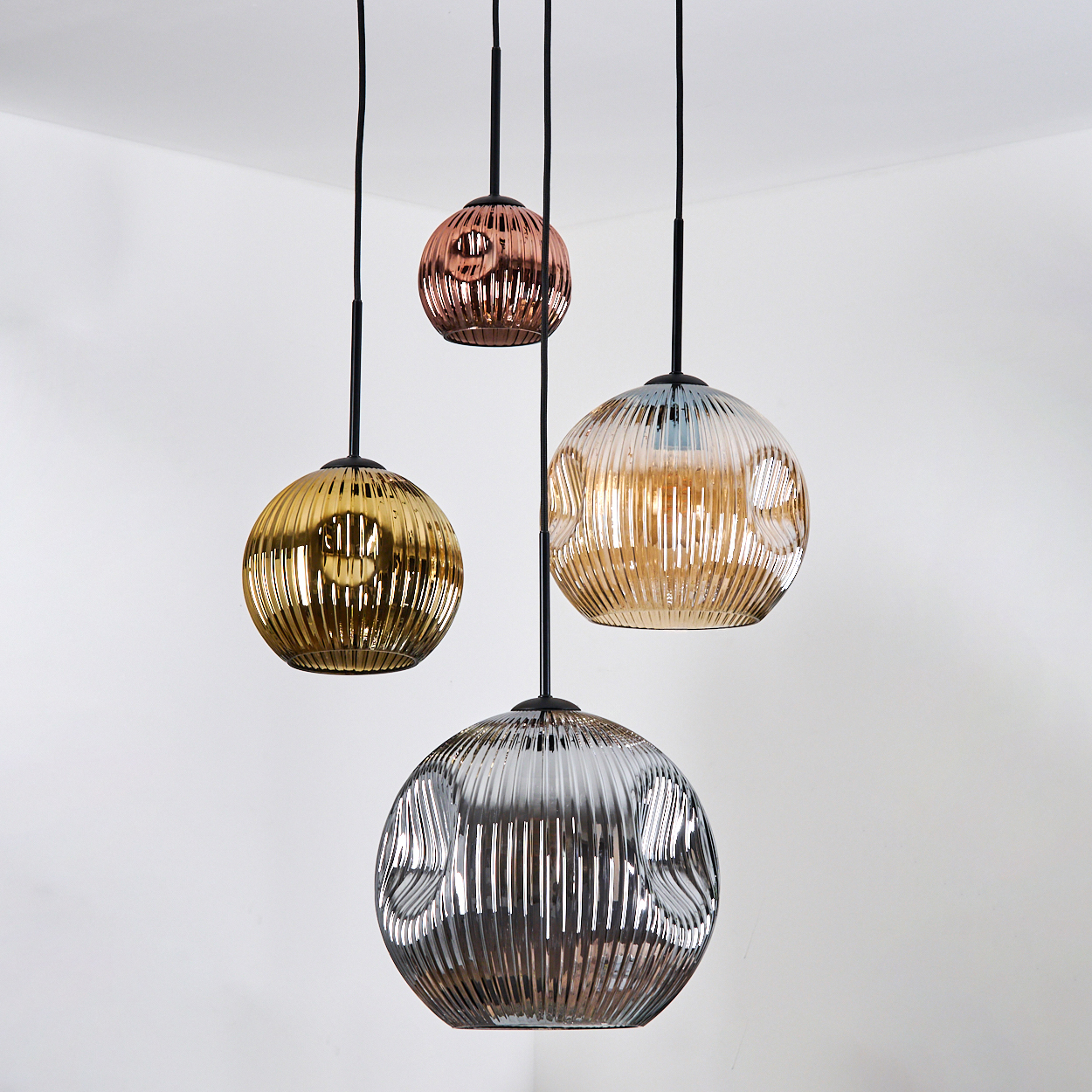 Vier kugelförmige Pendelleuchten in verschiedenen Farben: Gold, Kupfer, Klar und Silber, hängen an schwarzen Kabeln an einer weißen Wand.