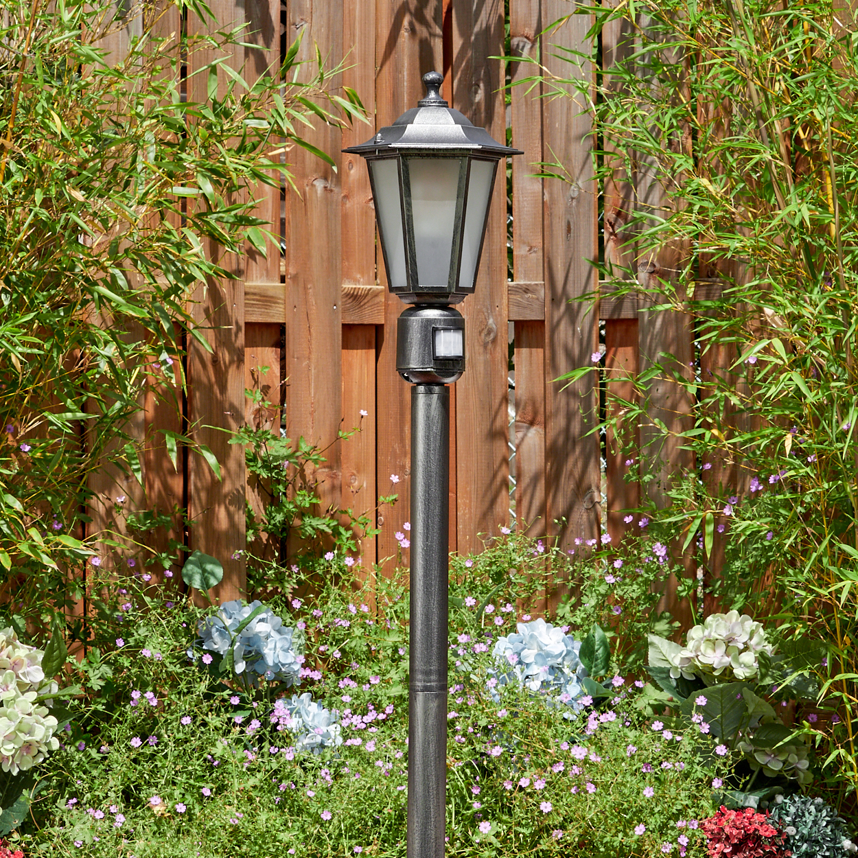 Eine Metall-Laterne steht in einem Garten mit Bambus, Blumen und einem Holzzaun im Hintergrund. Die Lampe hat einen Glasschirm.