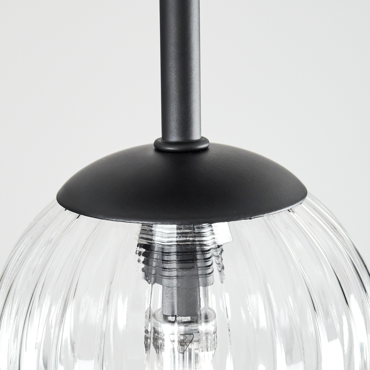 Nahaufnahme einer schwarzen Hängelampe. Sie hat einen Glasschirm mit vertikalen Rippen, eine schwarze Metallkappe und eine Metallstange.