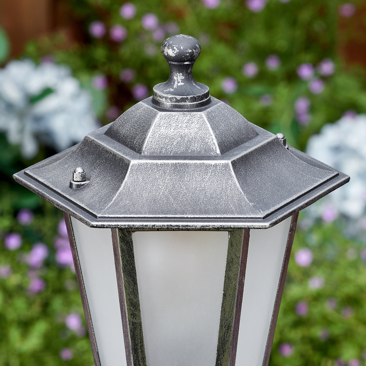 Nahaufnahme einer dekorativen Außenlampe mit silbernem Finish. Sie hat eine sechseckige Oberseite und Milchglasscheiben. Hintergrund mit Blumen.