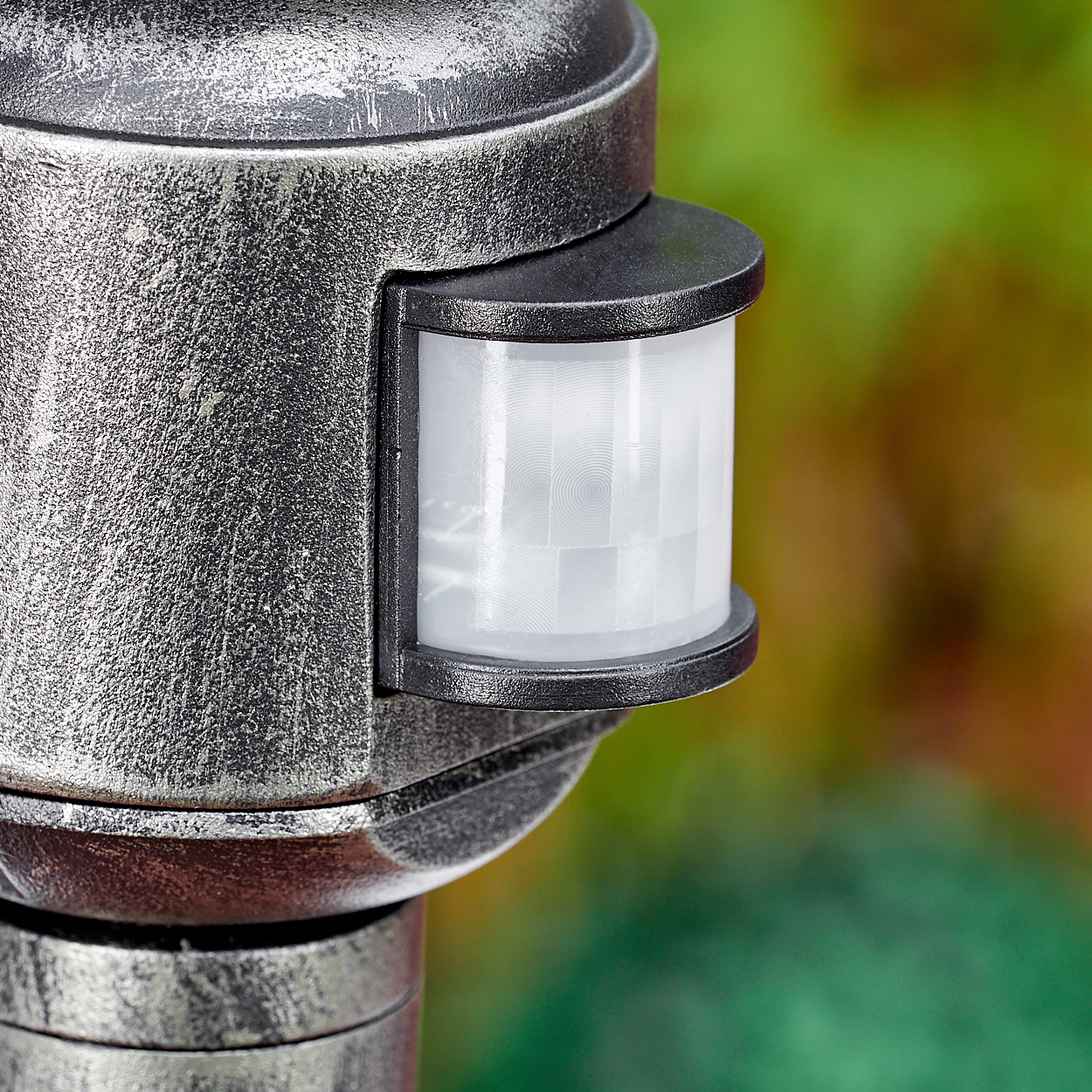 Nahaufnahme eines Bewegungssensors an einer metallischen Lampe. Der Sensor ist weiß und durch einen schwarzen Rahmen geschützt. Der Hintergrund ist verschwommen grün und braun.
