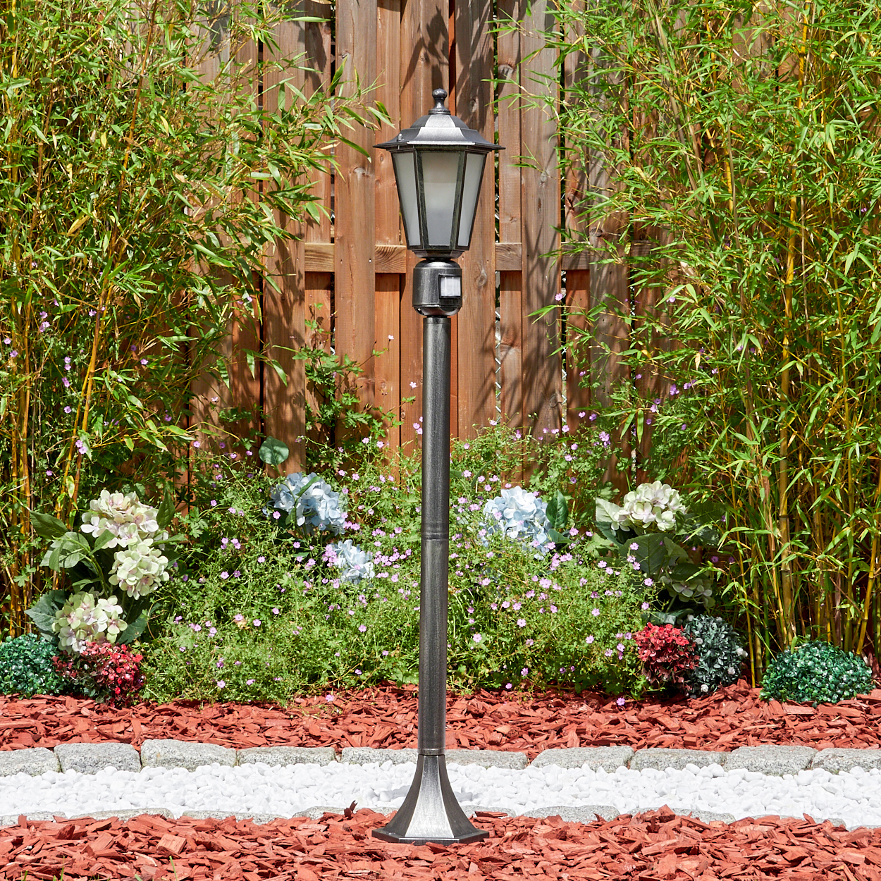 Eine dekorative Außenlampe steht in einem Garten. Bambus und ein Holzzaun befinden sich im Hintergrund. Blumen und Mulch umgeben sie.