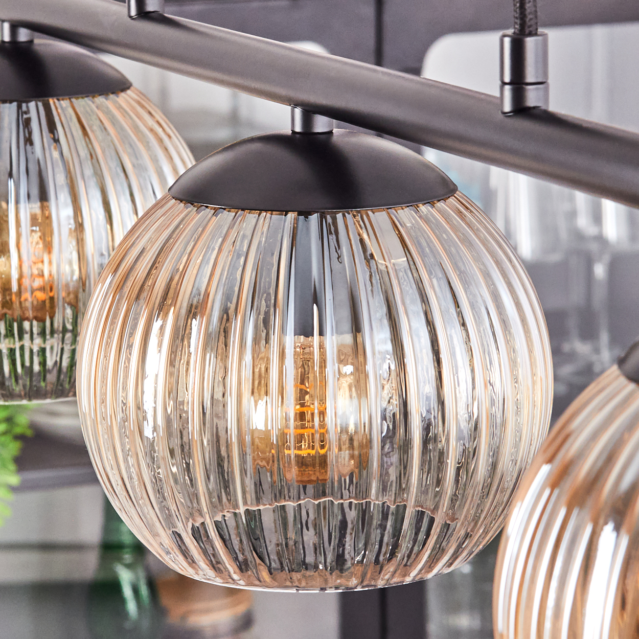 Nahaufnahme einer modernen Leuchte mit drei kugelförmigen, gerippten Glasschirmen, jeweils mit einer sichtbaren Glühbirne. Dunkler Metallrahmen.