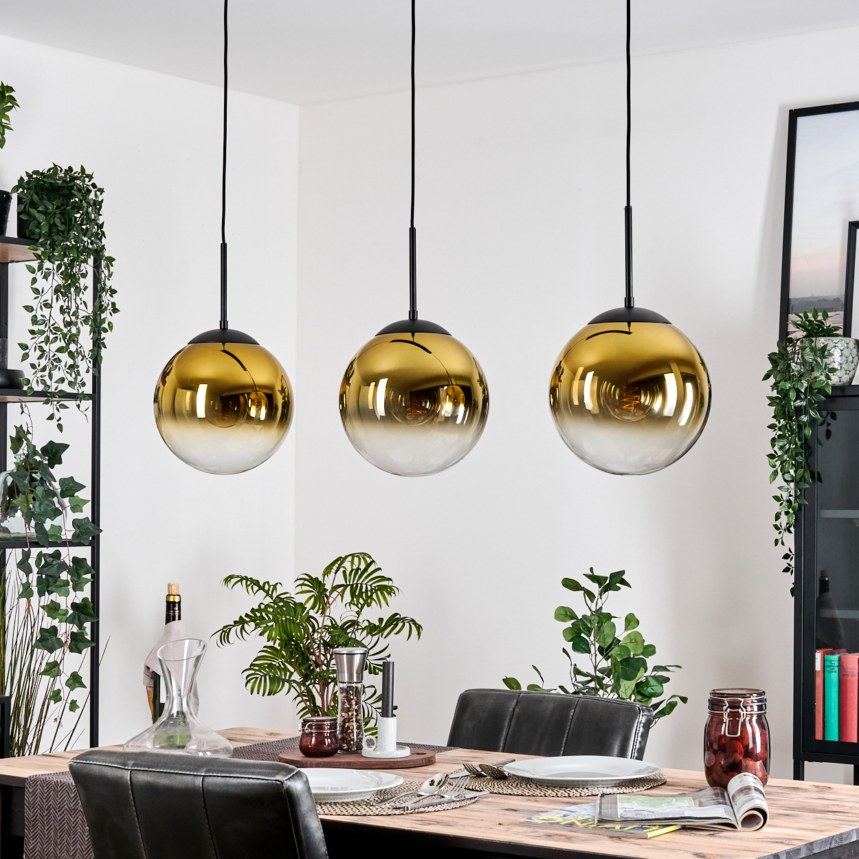 Drei goldene und gläserne Pendelleuchten hängen über einem Esstisch, der für das Abendessen gedeckt ist. Pflanzen und Dekorationsgegenstände umgeben den Tisch.