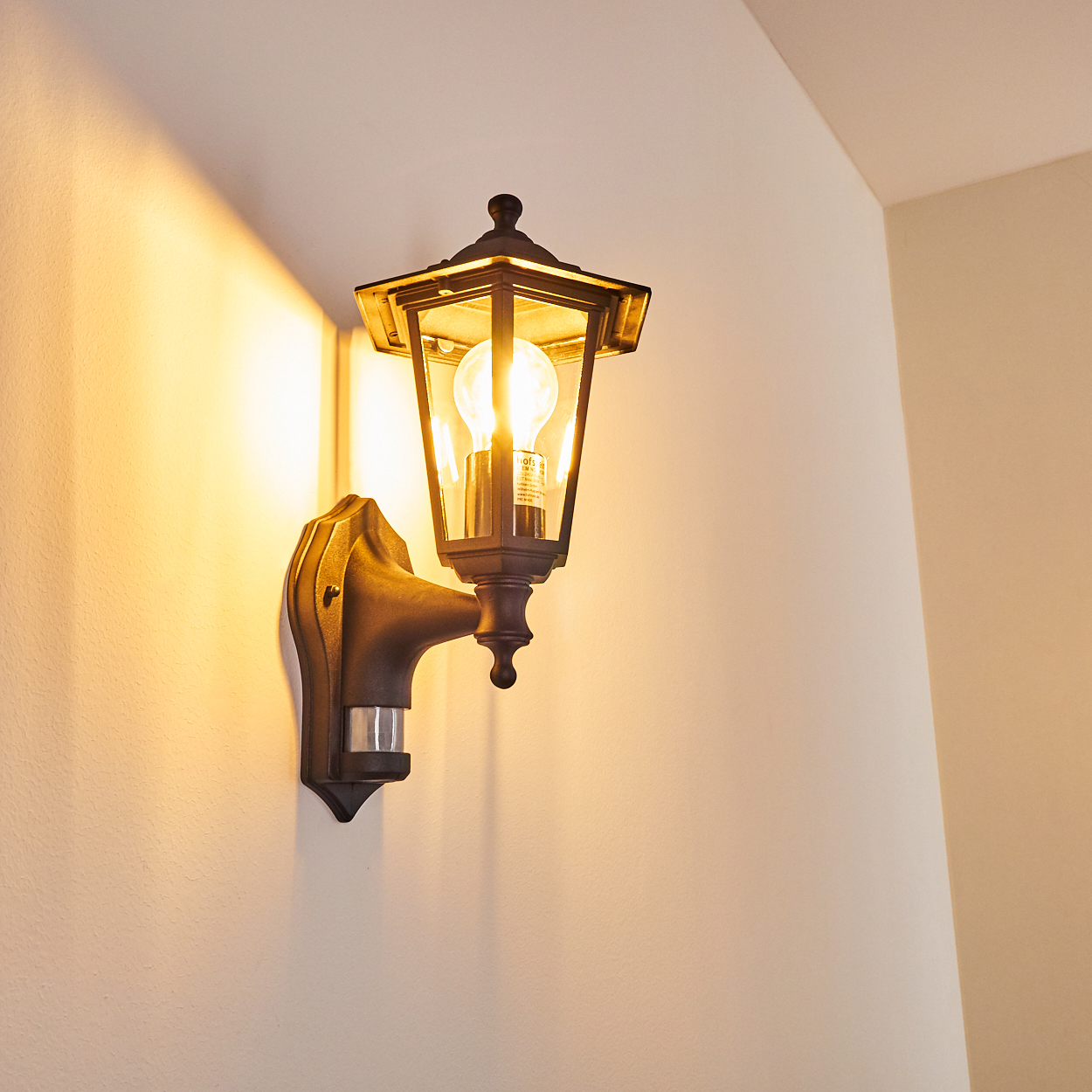 Eine dunkle Metall-Außenwandleuchte mit glühender Glühbirne ist an einer hellbeige Wand befestigt. Das Licht wirft Schatten.