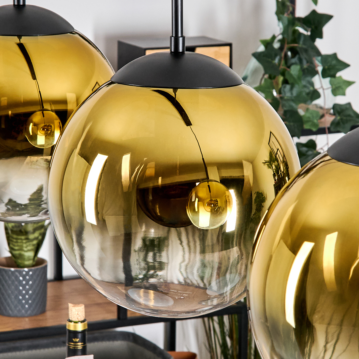 Drei goldene, kugelförmige Pendelleuchten hängen von der Decke. Eine Pflanze in einem grauen Topf steht darunter. Hintergrund unscharf.