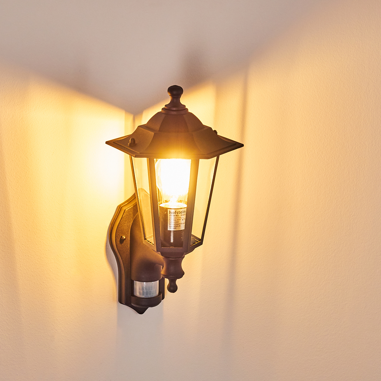 Dunkelbraune Außenlampe an hellbeige Wand. Die Lampe leuchtet, beleuchtet den Raum. Ein Bewegungssensor ist sichtbar.